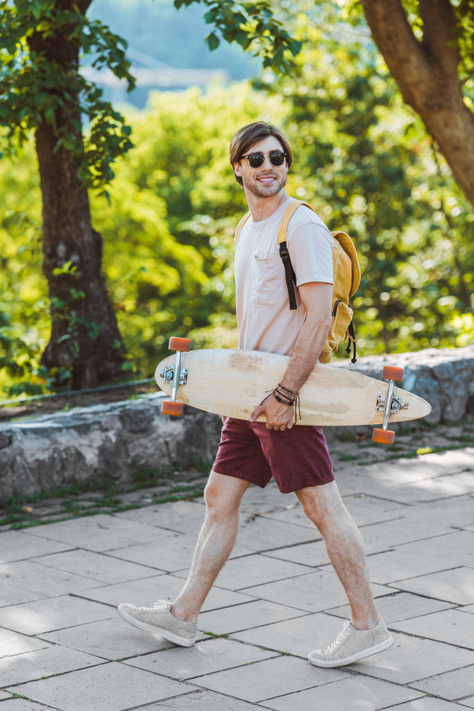 a napszemüveg az ember a hátizsák és a street longboard mosolyogva oldalnézete - Fotó, kép