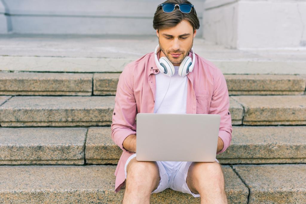 портрет человека с наушниками на шее с помощью ноутбука, сидя на ступеньках на улице
 - Фото, изображение