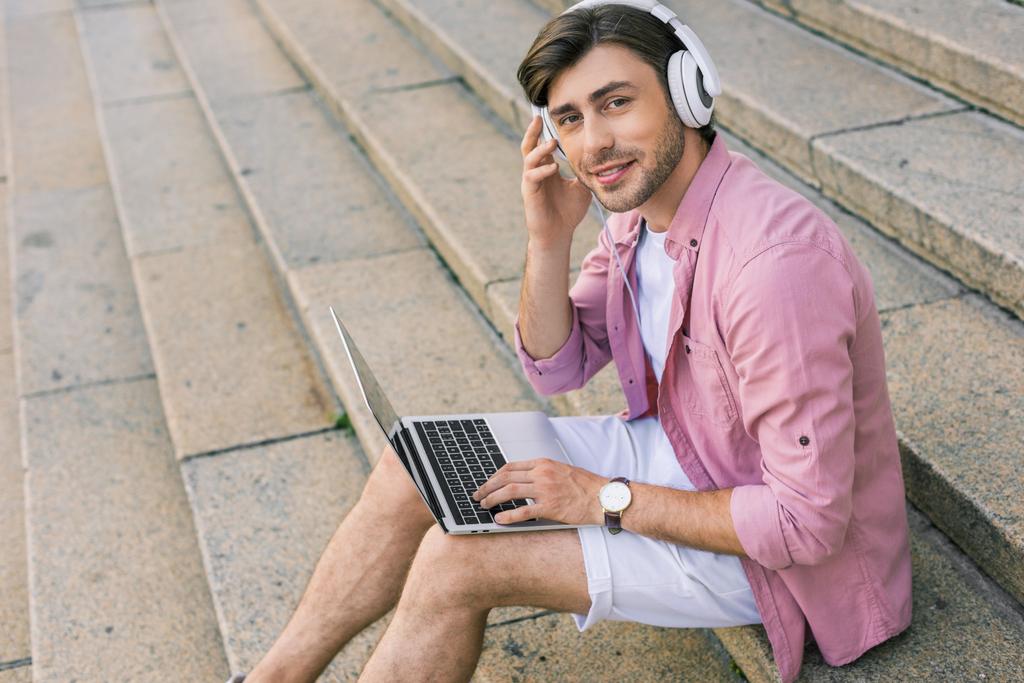 вид збоку стильного чоловіка в навушниках з ноутбуком, що сидить на сходах на вулиці
 - Фото, зображення