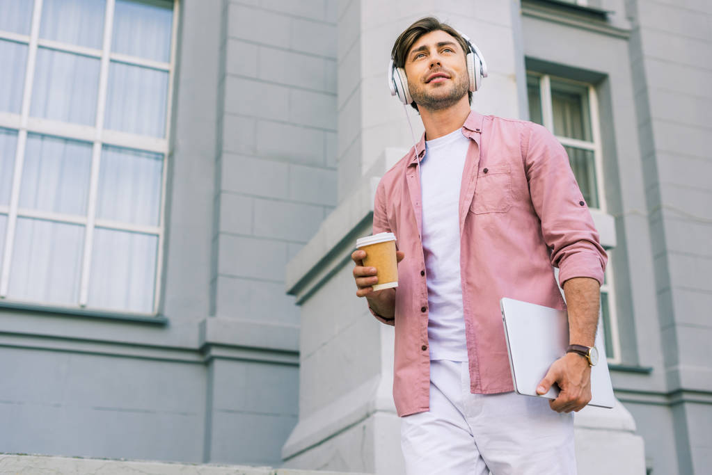 low angle view of young man in headphones with laptop and coffee to go on street - Photo, Image