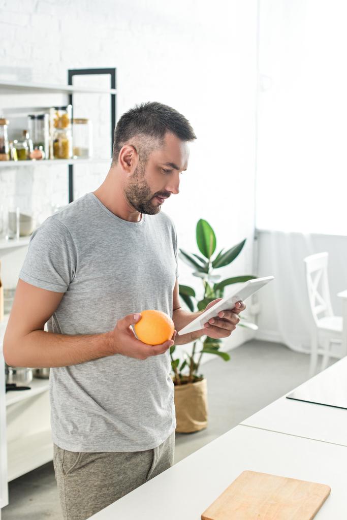 Πλάγια όψη του ανθρώπου που κρατώντας πορτοκαλί και ψάχνετε για συνταγή σε ψηφιακό tablet στην κουζίνα  - Φωτογραφία, εικόνα