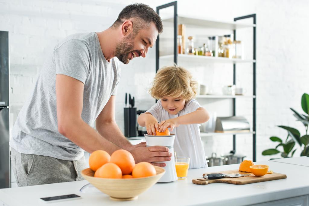 s úsměvem otce a malého syna, takže Čerstvý pomerančový džus podle ždímací na stole v kuchyni  - Fotografie, Obrázek