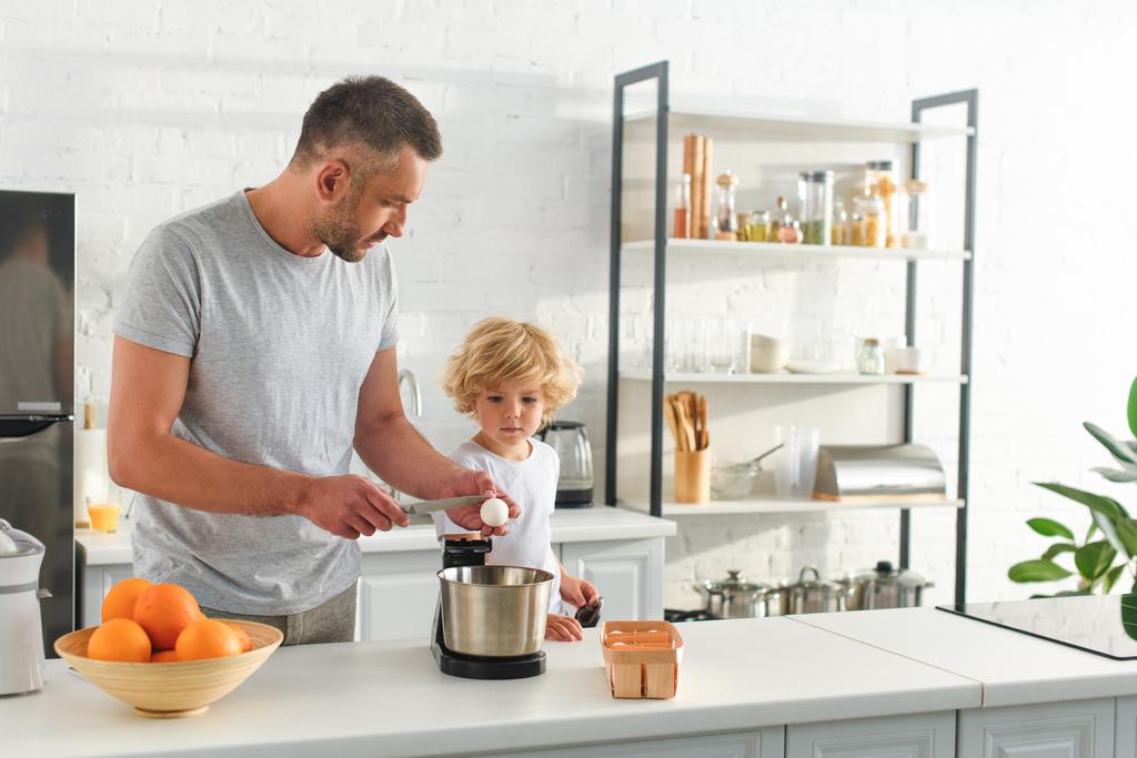 boční pohled člověka prolomit vejce nožem pro výrobu těsta a jeho syn stojící vedle v kuchyni  - Fotografie, Obrázek