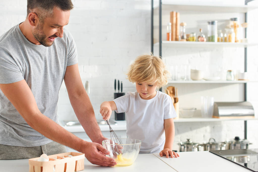 Отец держит миску, пока его сын взбивает яйца на кухне.
  - Фото, изображение