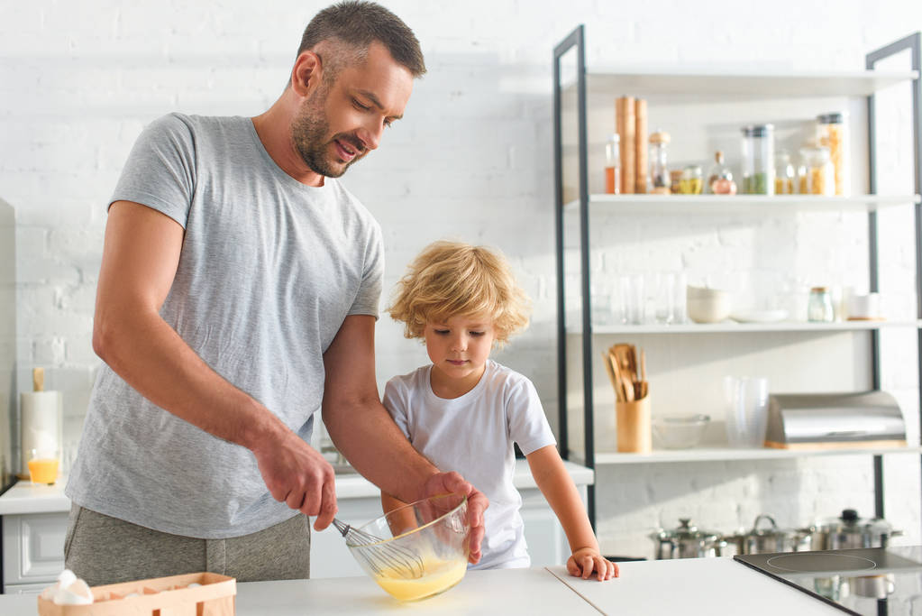 sonriente padre batiendo huevos en un tazón mientras su hijo de pie cerca en la cocina
  - Foto, Imagen