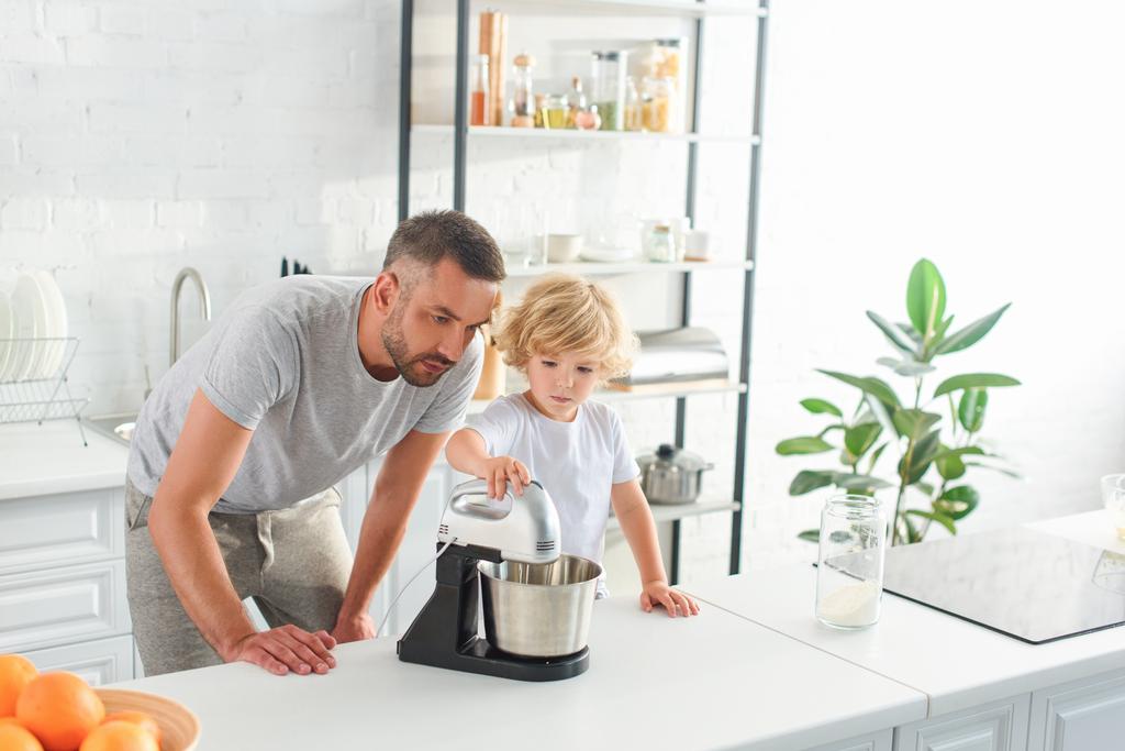 padre viendo cómo su hijo usando mezclador para hacer masa en la cocina
  - Foto, Imagen