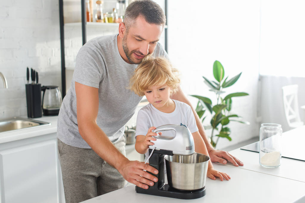 улыбающийся человек помогает сыну использовать миксер для приготовления теста на кухне
  - Фото, изображение