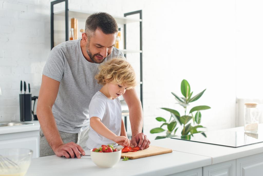 homem de pé com o filho enquanto ele tomando morango de tábua de corte na cozinha
 - Foto, Imagem