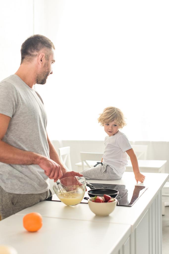vista lateral del padre batiendo masa en un tazón y hablando con su hijo mientras está sentado en la mesa en la cocina
 - Foto, Imagen