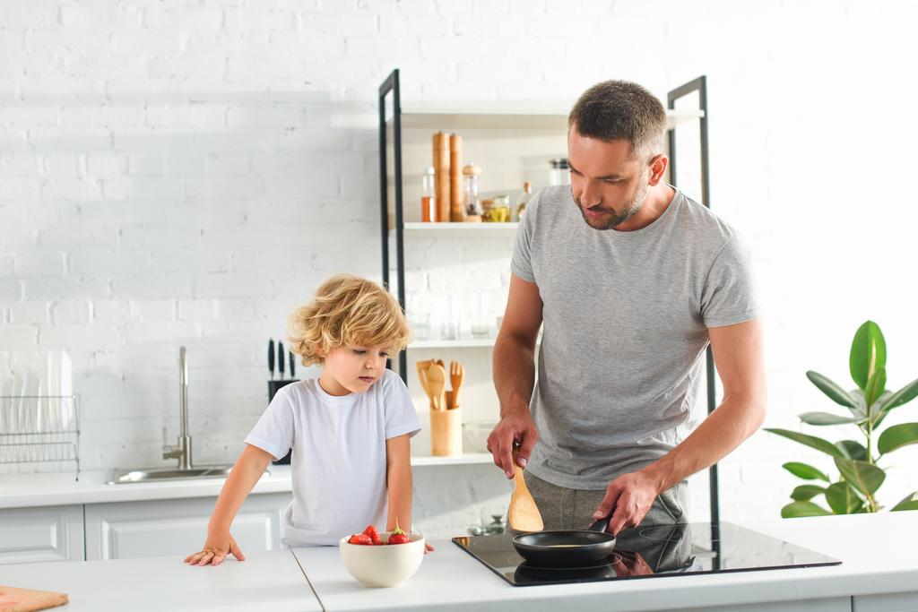 ο άνθρωπος θέρμανσης το τηγάνι ενώ ο γιος του στέκεται κοντά στην κουζίνα  - Φωτογραφία, εικόνα