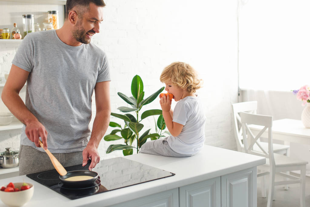 hombre haciendo panqueques en sartén mientras su hijo sentado cerca de la mesa en la cocina
 - Foto, Imagen