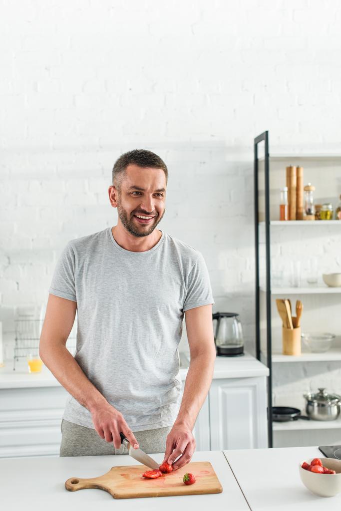 sorrindo homem adulto cortando morango por faca na mesa na cozinha
 - Foto, Imagem