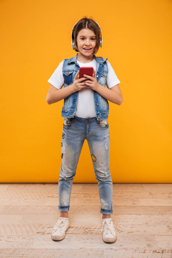 Full length portrait of a cute little schoolgirl listening to music with headphones while holding mobile phone over yellow background - Photo, Image