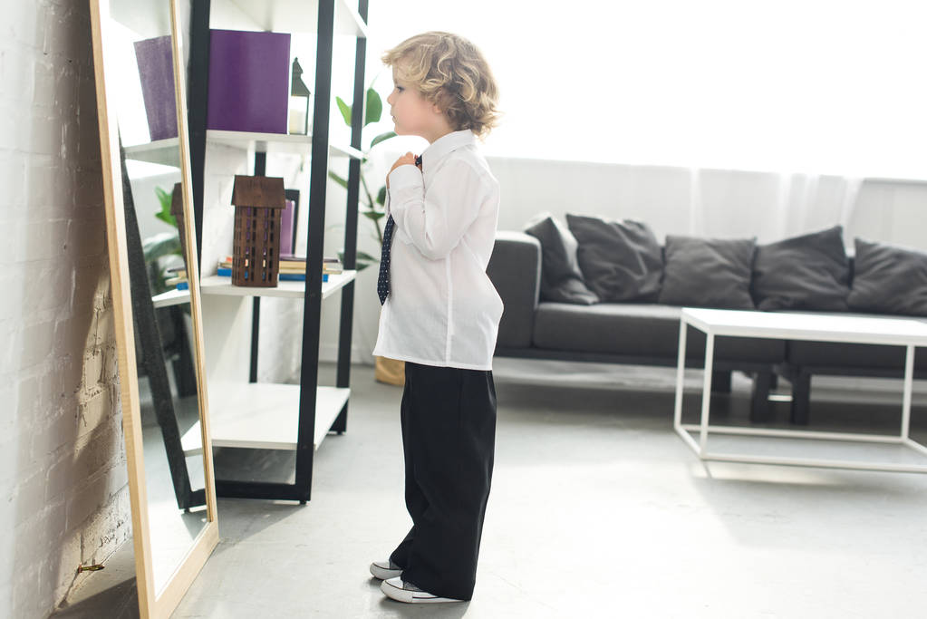 Zijaanzicht van de kleine jongen koppelverkoop stropdas over een wit overhemd voor spiegel thuis  - Foto, afbeelding
