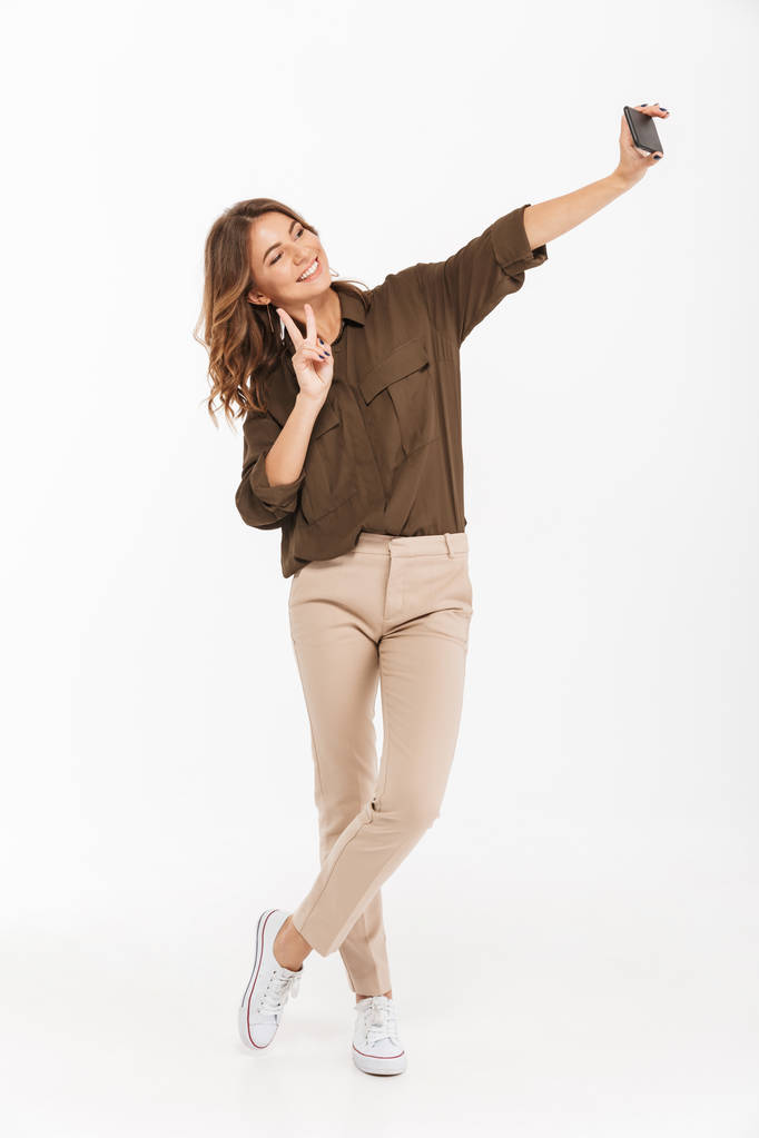 Retrato de larga duración de una joven sonriente tomando selfie con teléfono móvil aislado sobre fondo blanco
 - Foto, Imagen