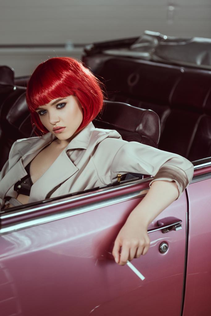 beautiful young woman in red wig holding cigarette and sitting in vintage car  - Photo, Image