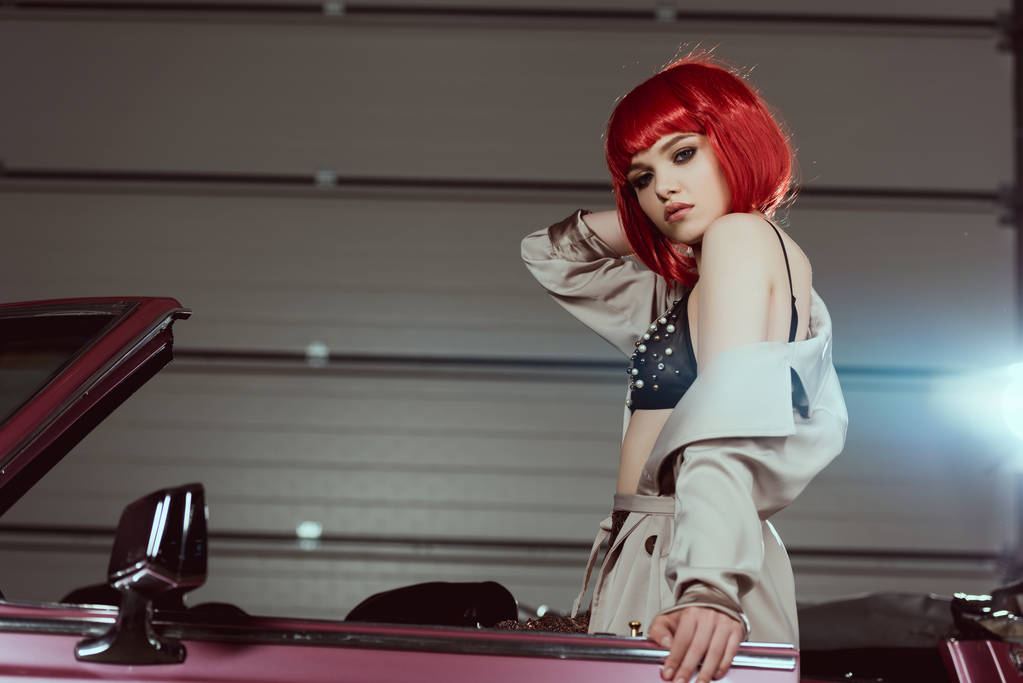с низким углом обзора стильной девушки в бюстгальтере и плаще, стоящей возле винтажного автомобиля и смотрящей в камеру
 - Фото, изображение