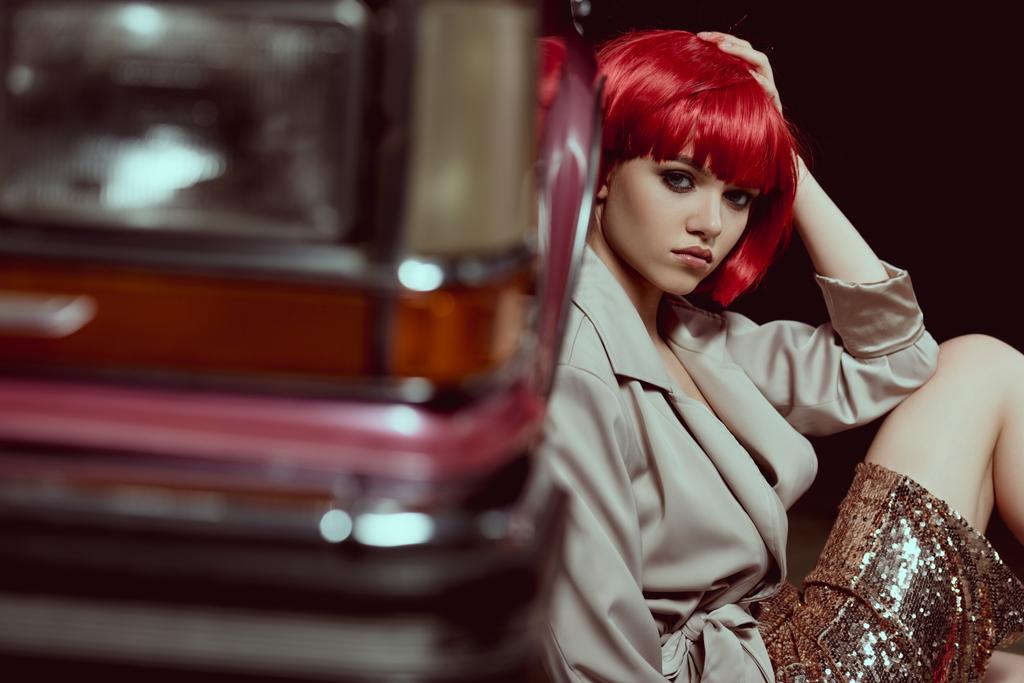selective focus of beautiful woman in red wig looking at camera while sitting near retro car - Photo, Image