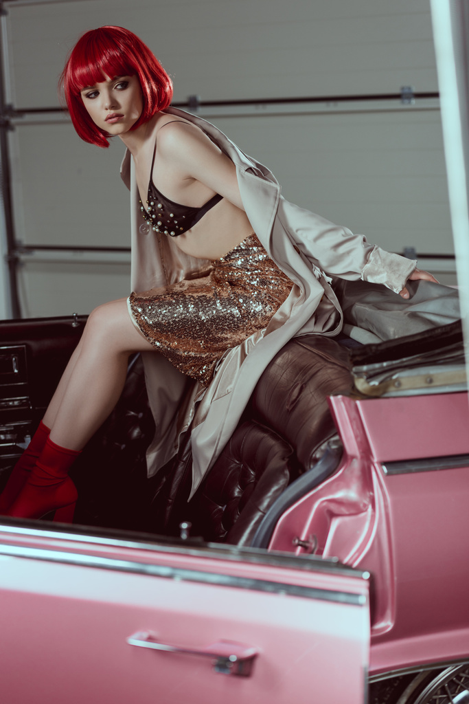 young woman in trench coat, glittering skirt and bra looking away while sitting on retro car - Photo, Image