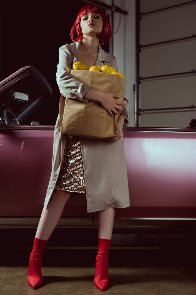 low angle view of beautiful blonde woman in red wig and stylish trench coat holding paper bag with lemons and looking at camera while standing near classic car - Photo, Image