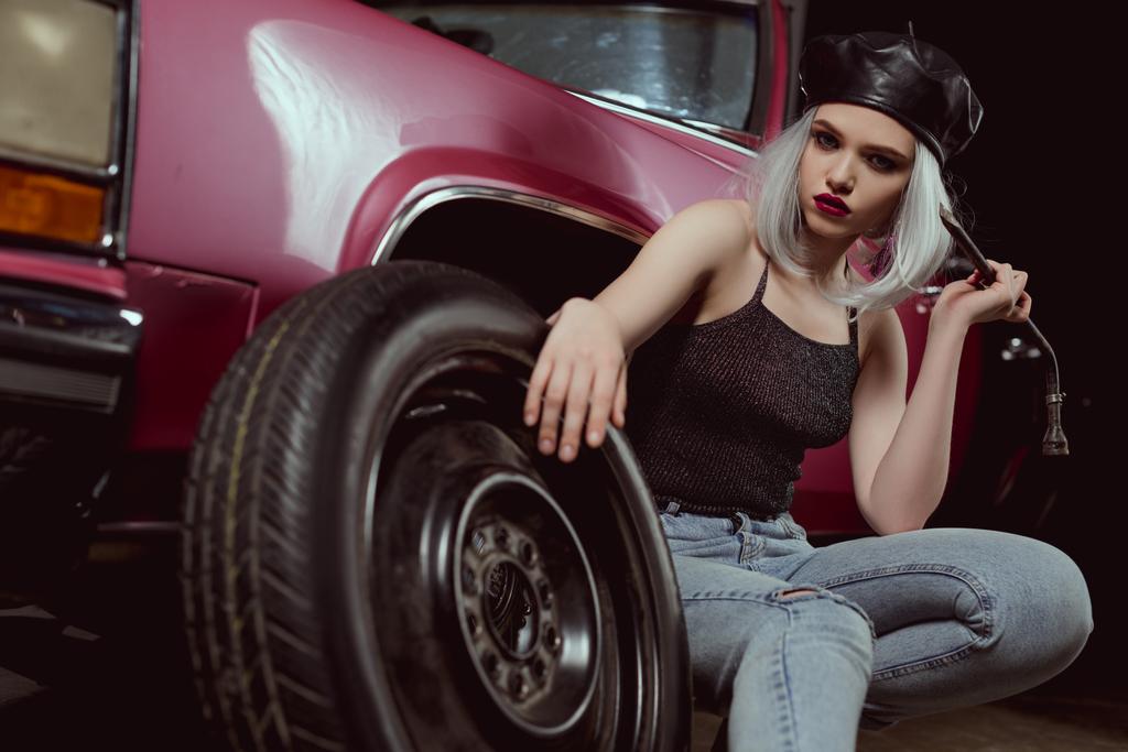 hermosa chica rubia con estilo mirando a la cámara mientras cambia la rueda del coche
 - Foto, Imagen