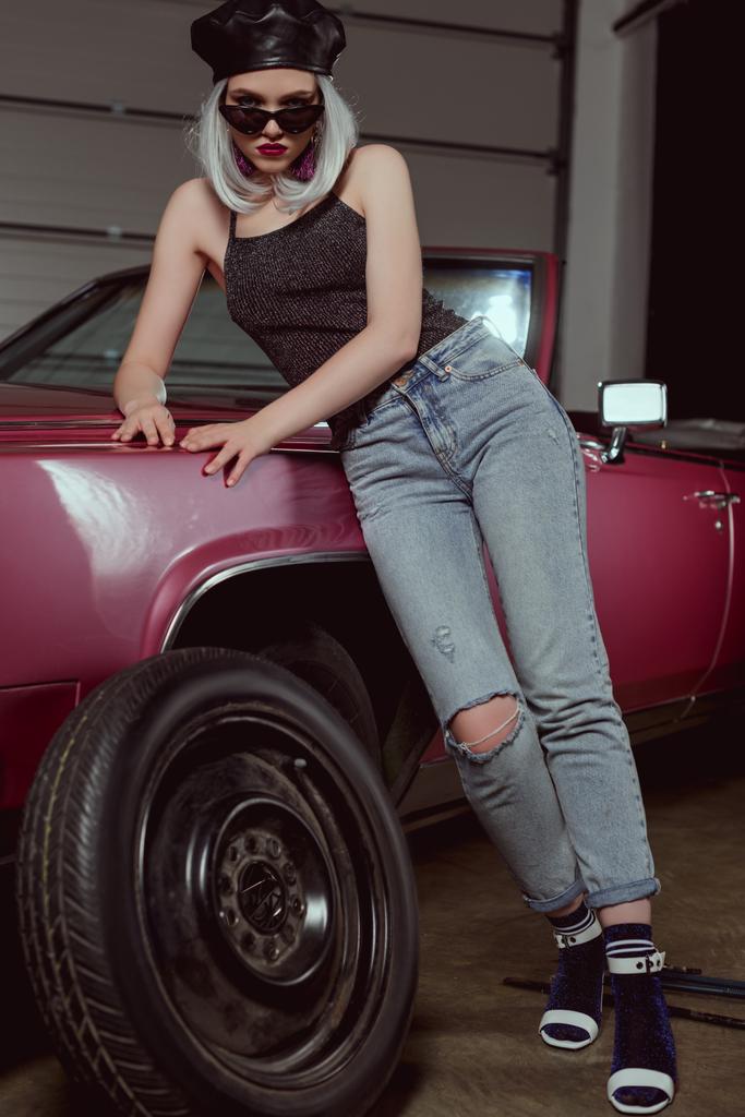 low angle view of stylish blonde girl in sunglasses and beret leaning at car and looking at camera - Photo, Image