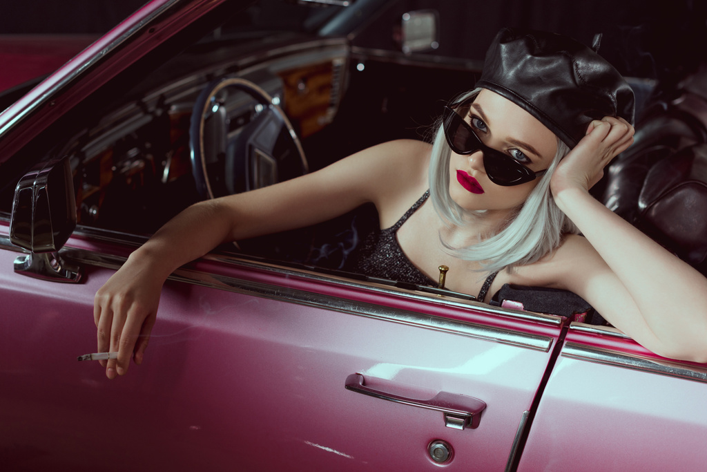 stylische blonde Frau mit Baskenmütze und Sonnenbrille, Zigarette in der Hand und Blick in die Kamera, während sie im Retro-Auto sitzt - Foto, Bild
