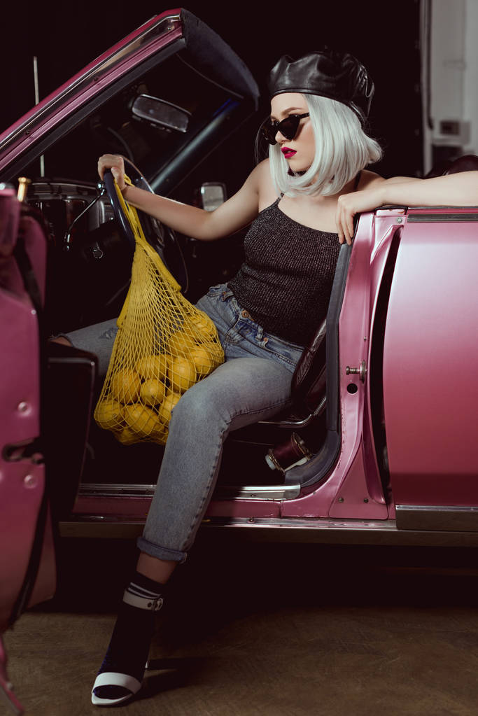 bella ragazza bionda elegante in berretto e occhiali da sole tenendo borsa stringa con limoni mentre seduto in auto retrò
   - Foto, immagini