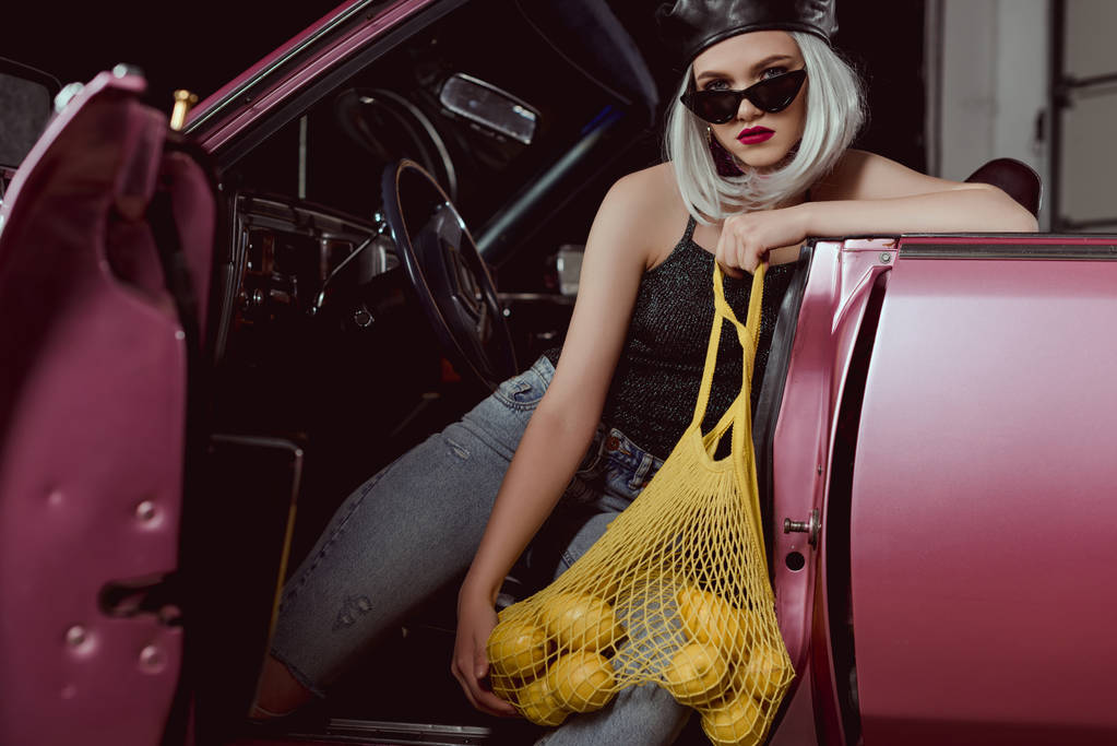 stylish blonde girl in beret and sunglasses holding string bag with lemons while sitting in car   - Photo, Image