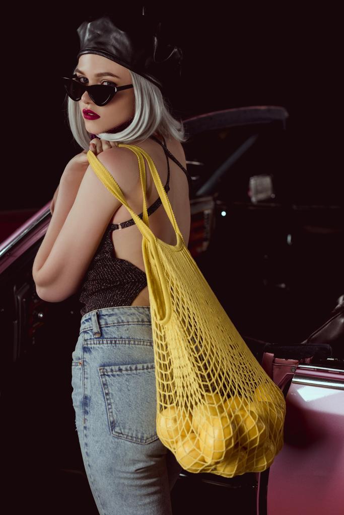stylowa dziewczyna Blondynka w okularach słonecznych i beret trzymając worek ciąg z cytryn, stojący w pobliżu zabytkowych samochodów    - Zdjęcie, obraz