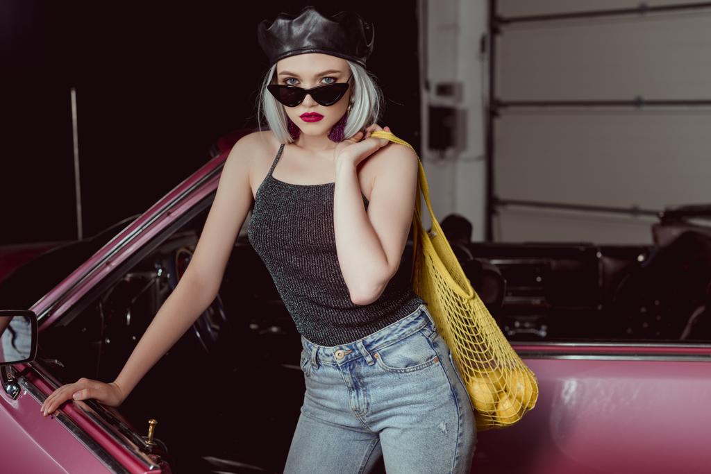 elegante chica rubia en gafas de sol y boina sosteniendo bolsa de cuerda con limones y mirando a la cámara mientras está de pie cerca del coche
    - Foto, Imagen