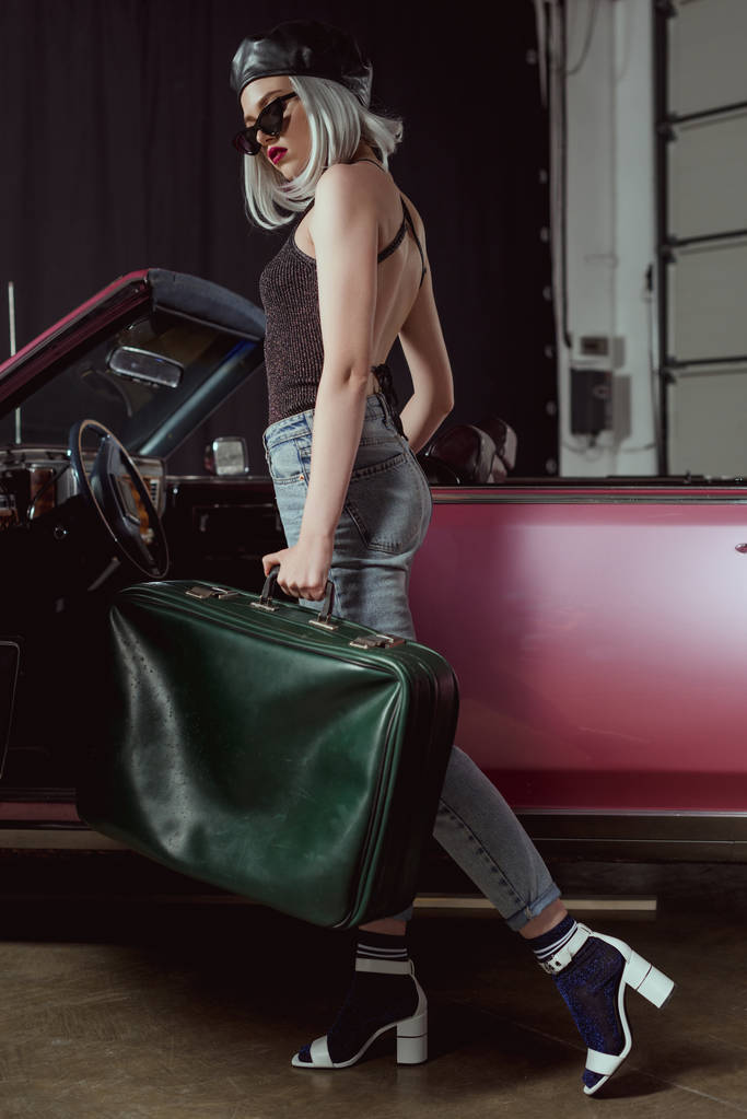 полный вид на стильную блондинку в солнечных очках и берете, держащую старомодный ретро-автомобиль с чемоданом
 - Фото, изображение