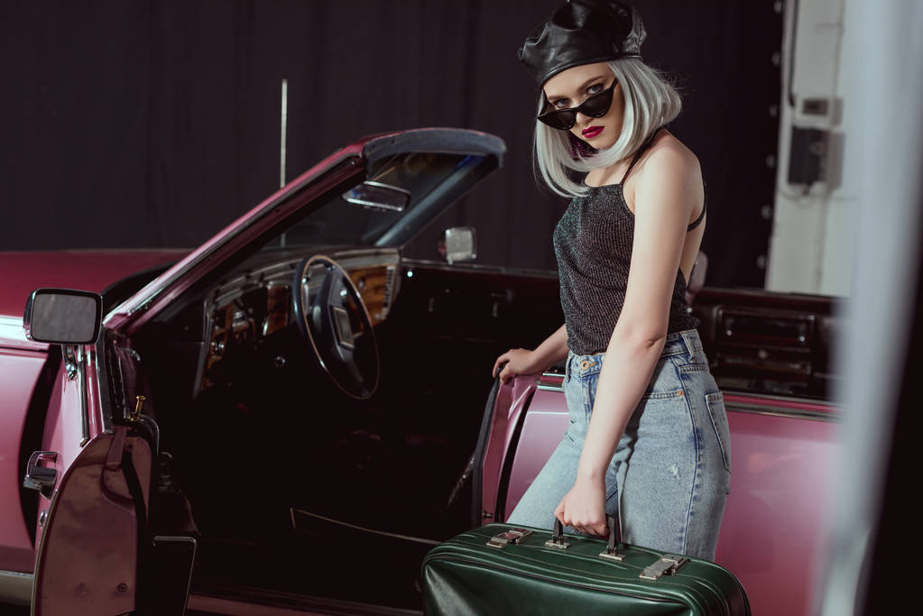 elegante chica rubia en gafas de sol y boina sosteniendo maleta retro y mirando a la cámara mientras está de pie cerca de coche marrón
  - Foto, Imagen