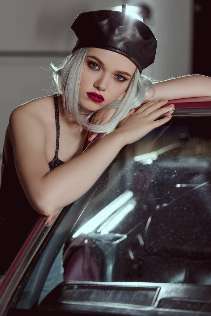 kaunis tyylikäs blondi nainen musta nahka baskeri nojaa autoon ja katselee kameraa
 - Valokuva, kuva