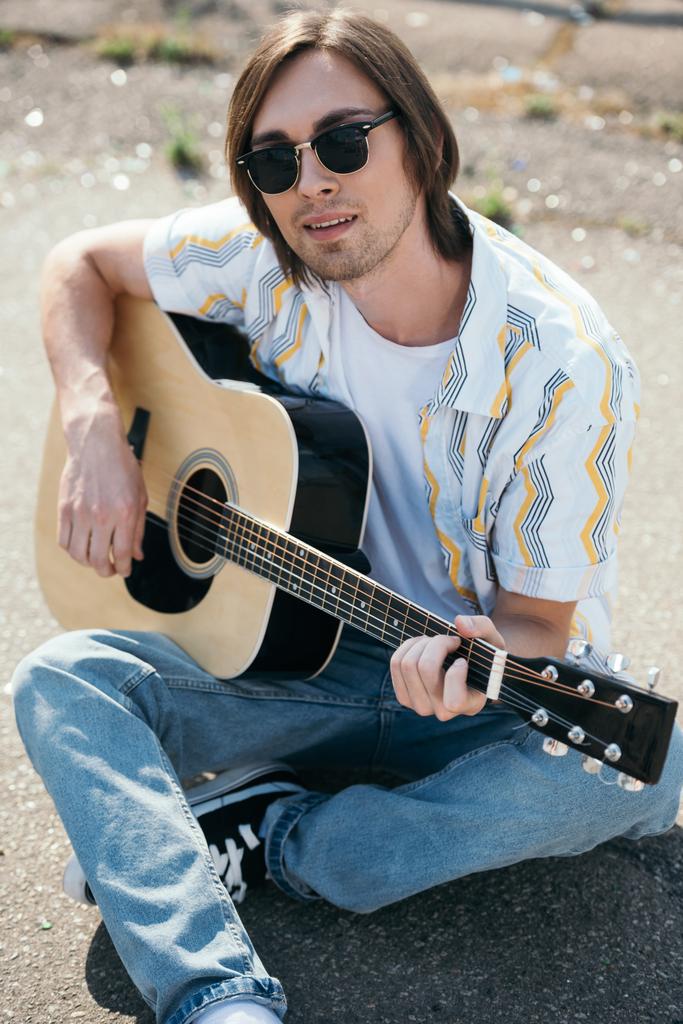 Νεαρός παίζει κιθάρα και να κάθεται στο έδαφος σε αστικό περιβάλλον - Φωτογραφία, εικόνα