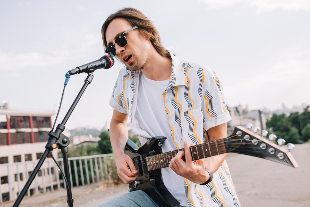 Young man in sunglasses playing guitar and singing in urban environment - Photo, Image