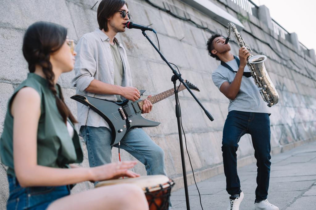 Уличные музыканты выступают с гитарой, барабаном и саксофоном на солнечной улице города
 - Фото, изображение