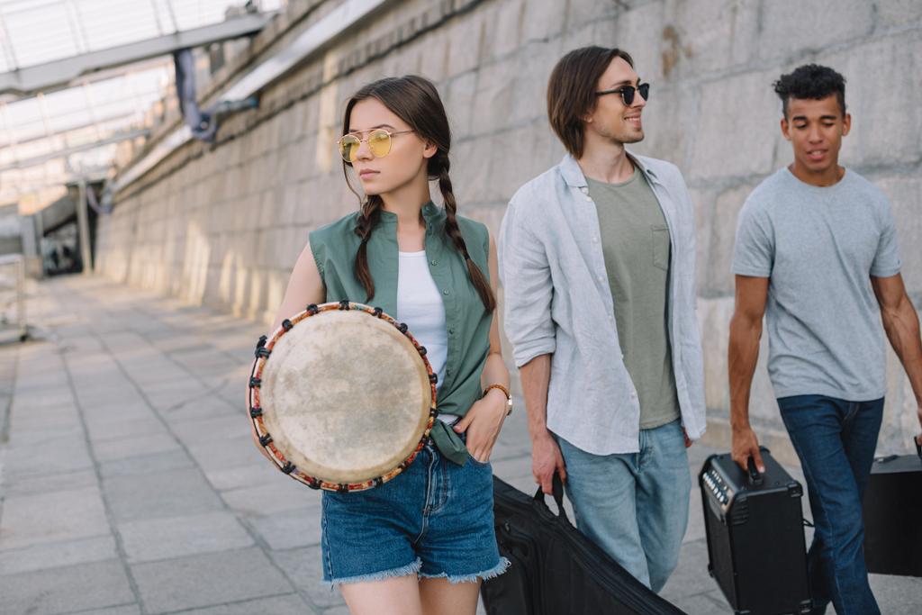 Equipe de jovens amigos do sexo masculino e feminino caminhando e carregando instrumentos musicais em ambiente urbano
 - Foto, Imagem