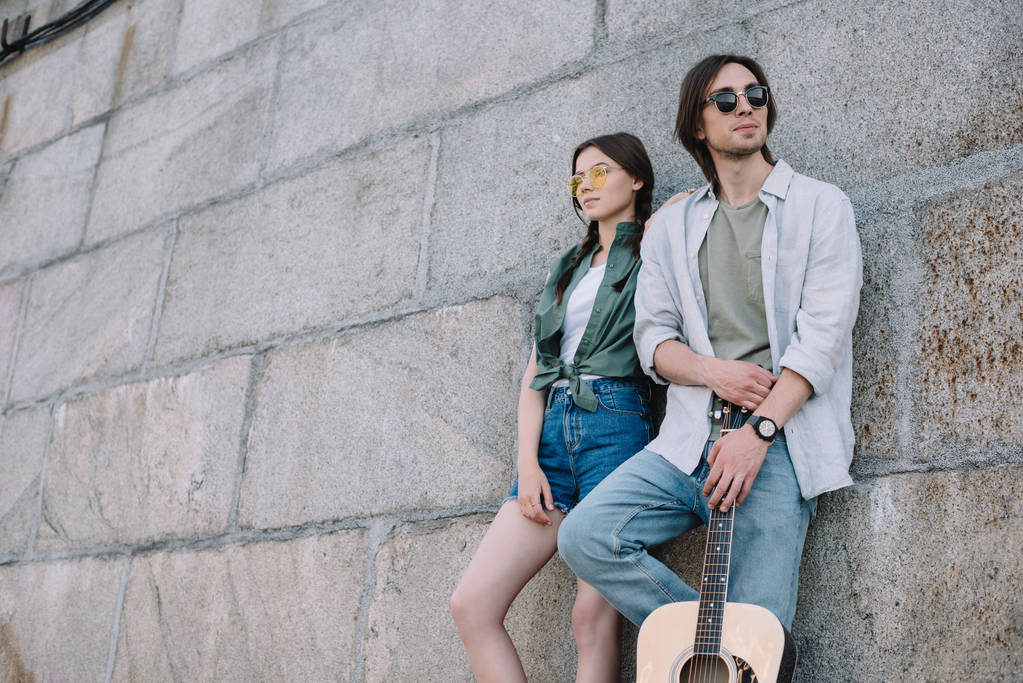 Молодая девушка и мужчина в солнечных очках держат гитару за стеной на улице
 - Фото, изображение