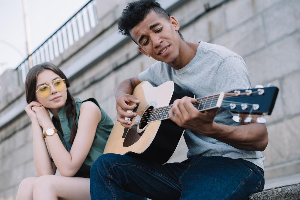 Menina bonita ouvindo o homem afro-americano tocando guitarra em ambiente urbano
 - Foto, Imagem