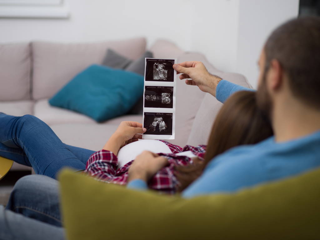 Νεαρή έγκυος ζευγάρι ψάχνει φωτογραφία υπερηχογράφημα του μωρού ενώ χαλαρώνετε στον καναπέ στο σπίτι - Φωτογραφία, εικόνα