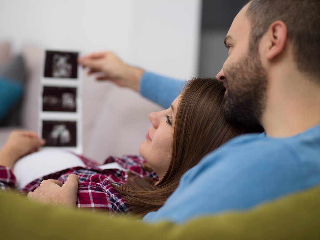 Νεαρή έγκυος ζευγάρι ψάχνει φωτογραφία υπερηχογράφημα του μωρού ενώ χαλαρώνετε στον καναπέ στο σπίτι - Φωτογραφία, εικόνα