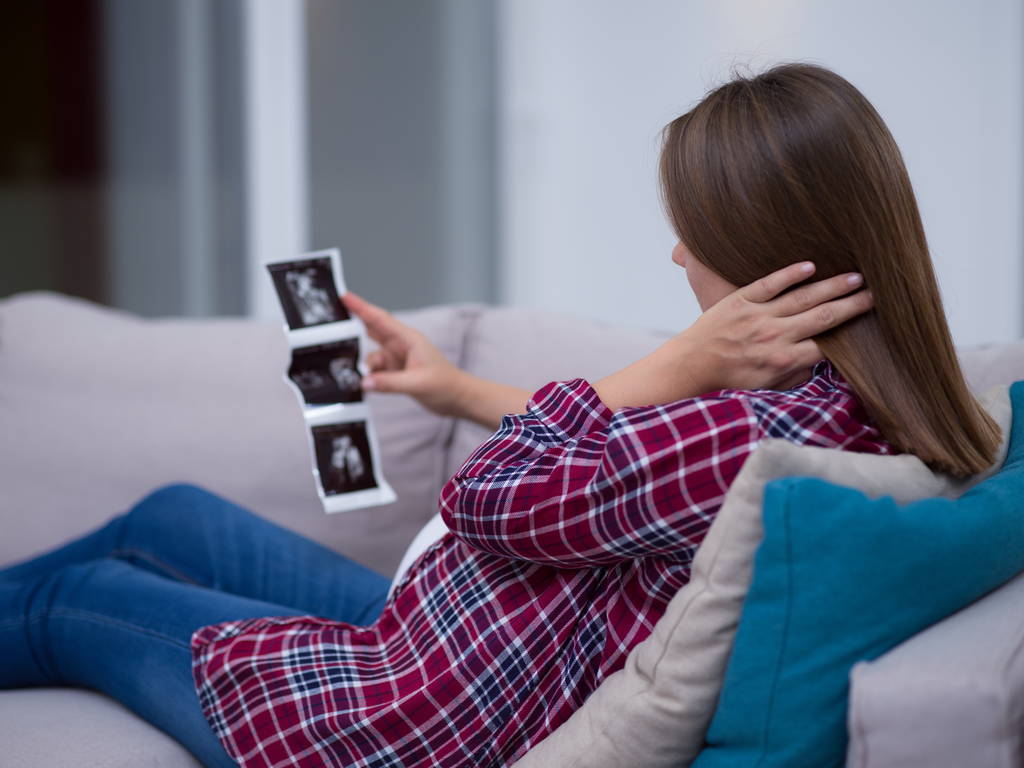 junge schwangere Frau sucht Baby-Ultraschall-Foto, während sie es sich zu Hause auf dem Sofa gemütlich macht - Foto, Bild