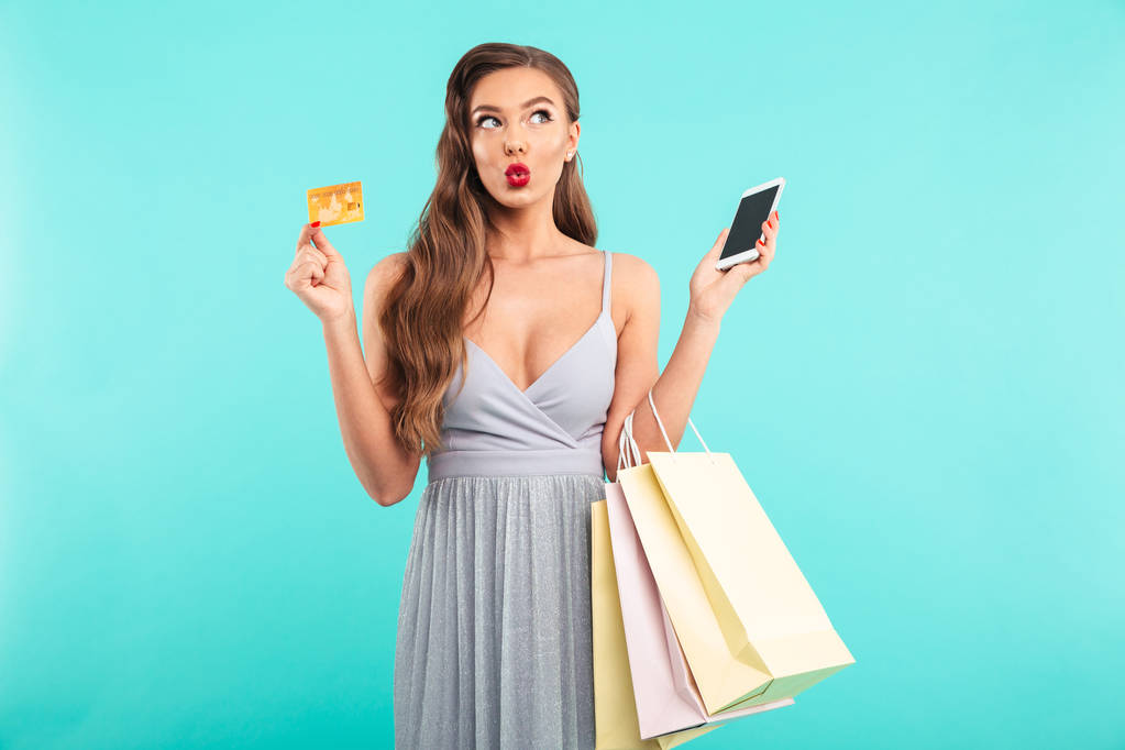 Очаровательная шопоголичка, держащая сумки для покупок и платящая смартфоном и кредитной картой на синем фоне
 - Фото, изображение