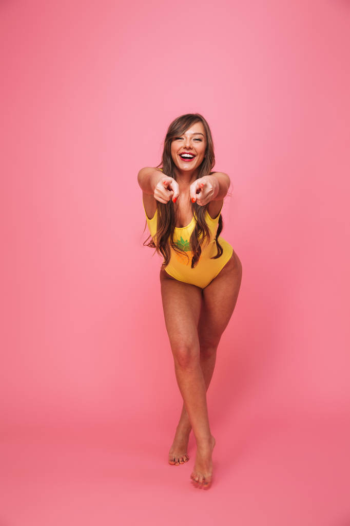 Full length portrait of a smiling young woman dressed in swimsuit pointing fingers at camera isolated over pink background - Photo, Image