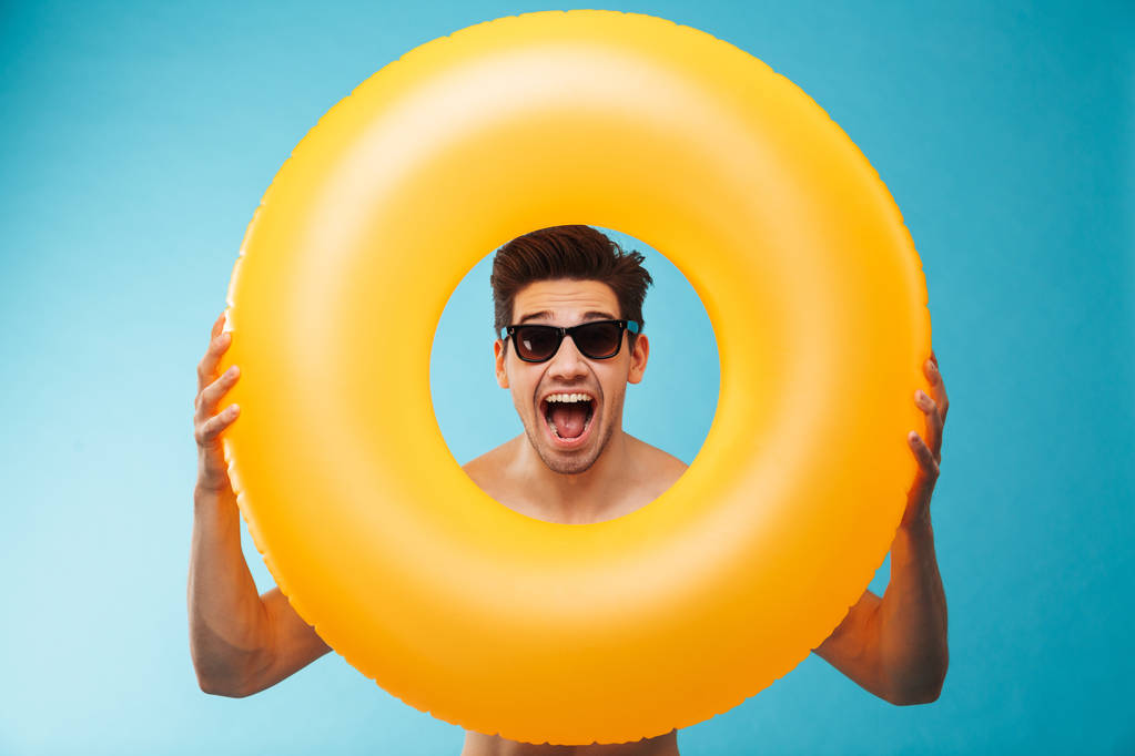 Close up of a cheerful man in sunglasses looking through inflatable ring at camera over blue background - Photo, Image