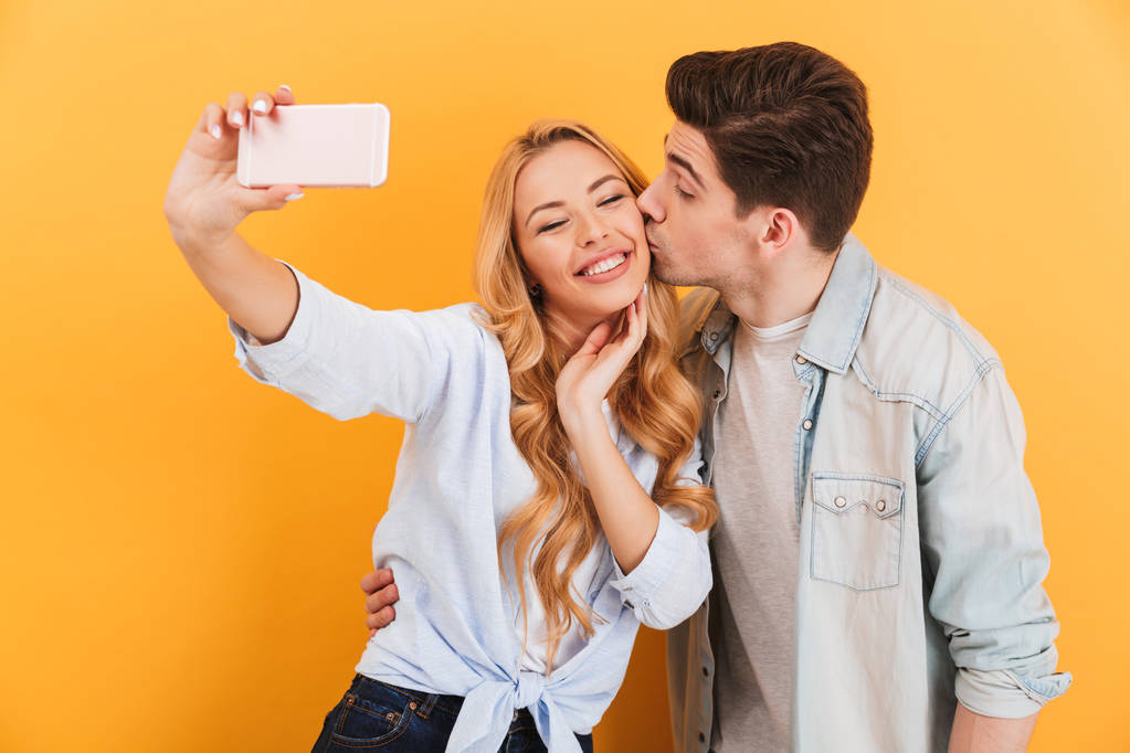 Porträt eines schönen Paares, das Selfie-Foto auf dem Handy macht, während Mann Frau auf Wange küsst, isoliert auf gelbem Hintergrund - Foto, Bild