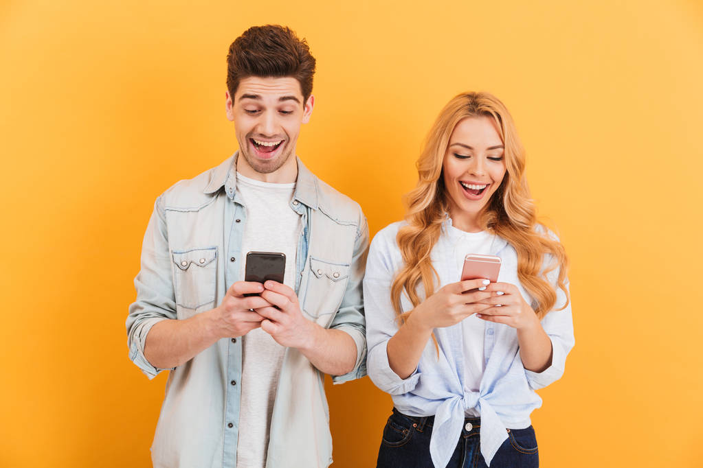 Foto van opgewonden mensen man en vrouw lachende tijdje beide gebruikend celtelefoons geïsoleerd op gele achtergrond - Foto, afbeelding