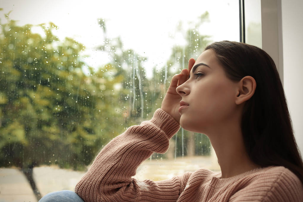 Nuori surullinen nainen istuu lähellä ikkunaa kotona
 - Valokuva, kuva