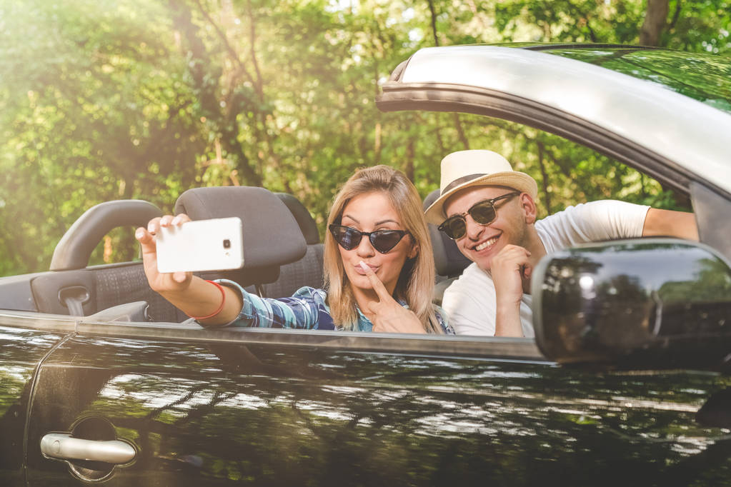 Casal jovem tirando selfie com smartphone enquanto sentado em carro conversível. Conceitos de viagens e tecnologia
. - Foto, Imagem